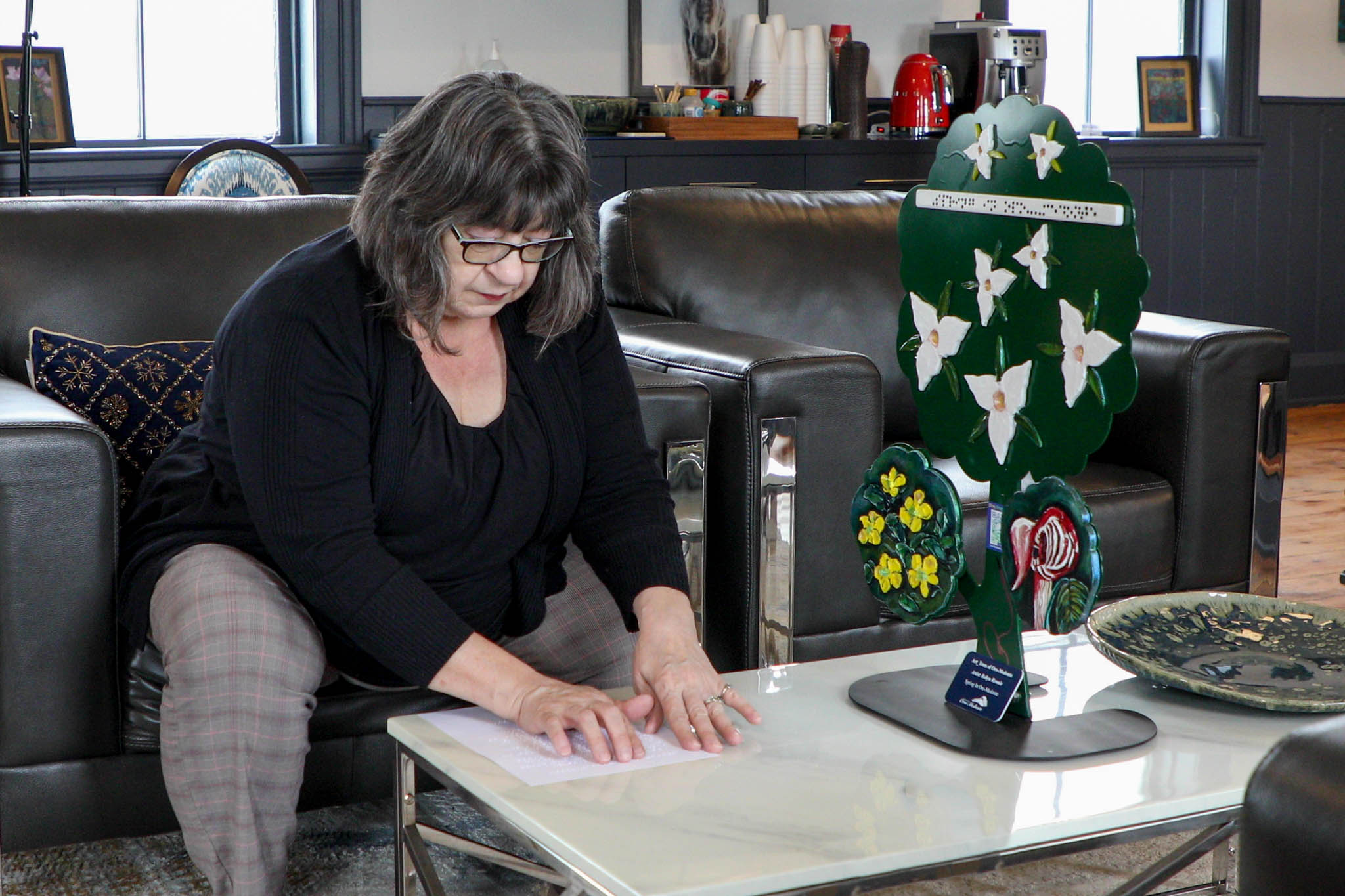 A woman with dark hair and glasses in a black top and grey pants reads a braille description of a sculpture on the table in front of her.